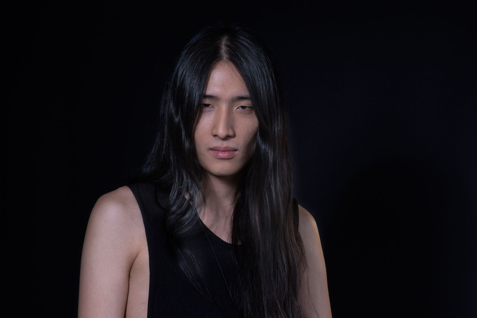 A person with long, dark hair stands against a solid black background. They are wearing a sleeveless black top and looking directly at the camera. The lighting is focused on the person, leaving the background dark and undefined. The individual’s expression is neutral or slightly serious.