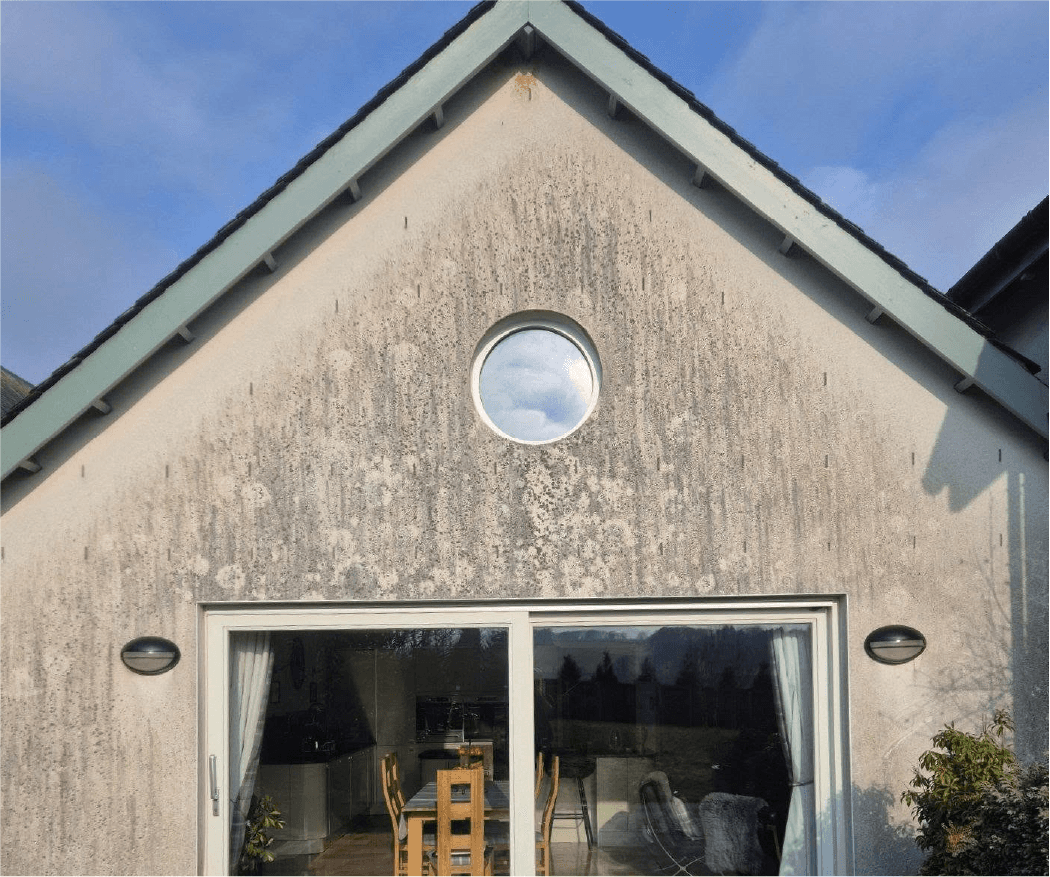 A triangular house facade with textured, weathered walls features a round window with a sky reflection and a sliding glass door.