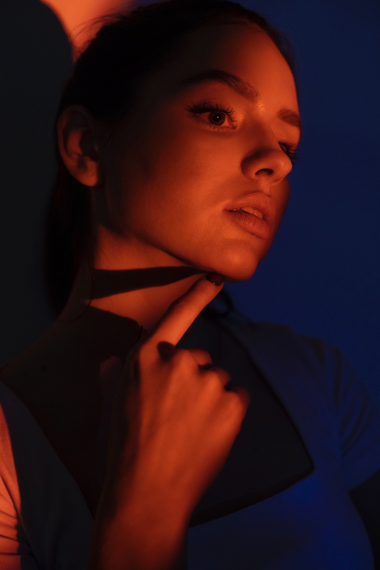 Good-looking young woman in front of a dark blue background, with orange lighting reflecting on her face, touching her chin with her index finger.