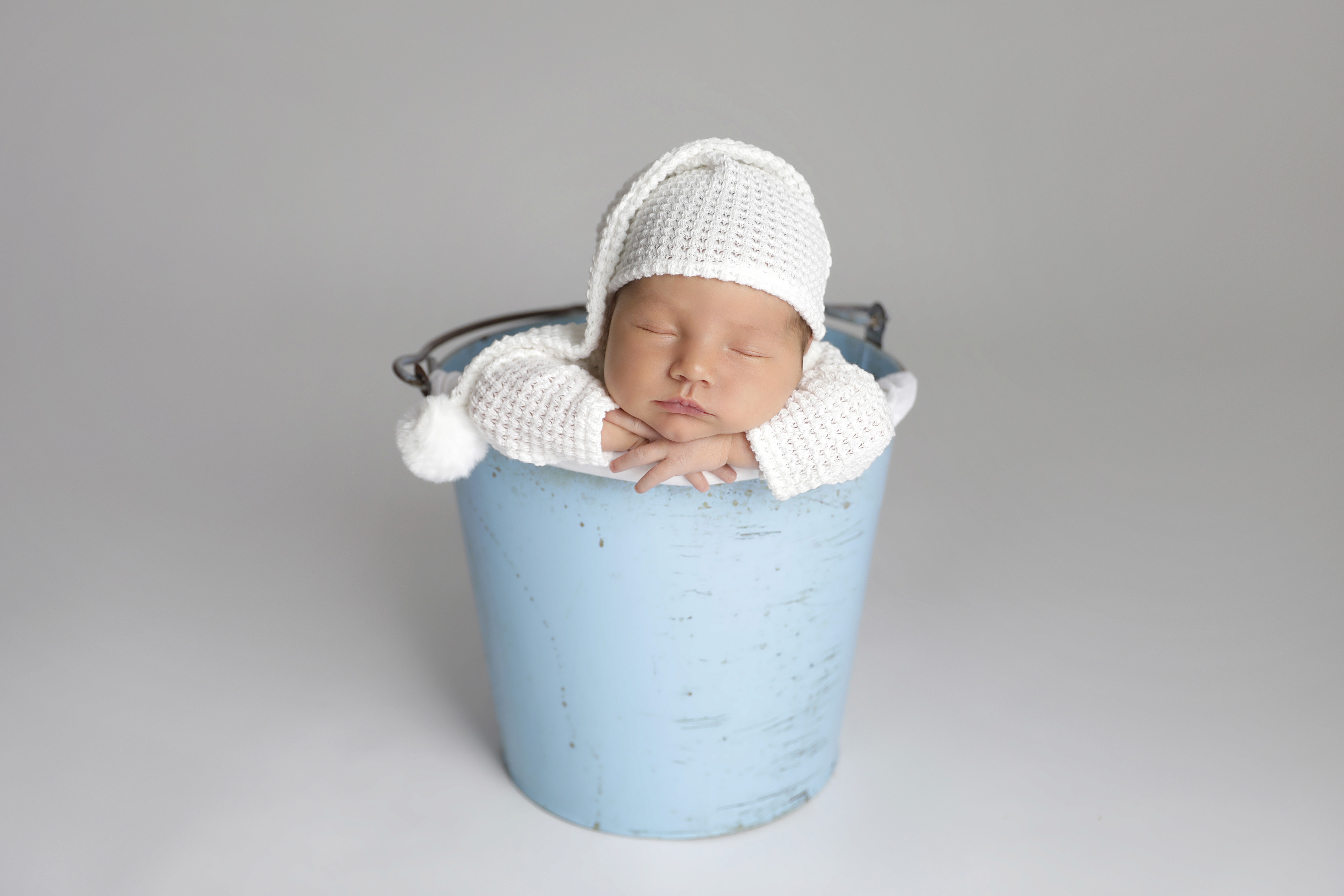 A baby in a white outfit is sleeping in a blue bucket amidst the grey background.&nbsp;