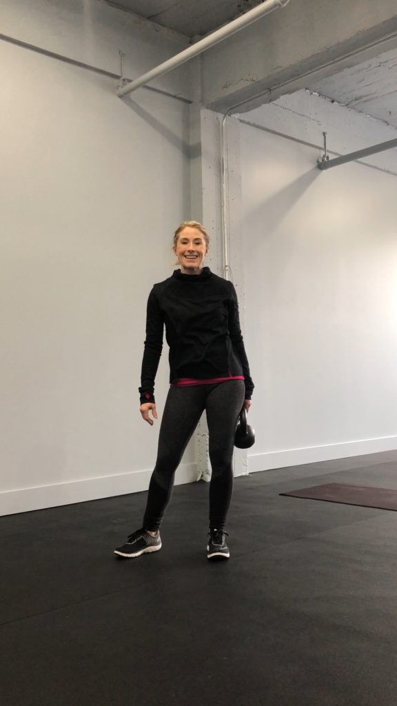 Heather Hausman in athletic wear stands in a minimalist gym setting holding a kettlebell, with gray walls and black flooring, embodying fitness and strength training.
