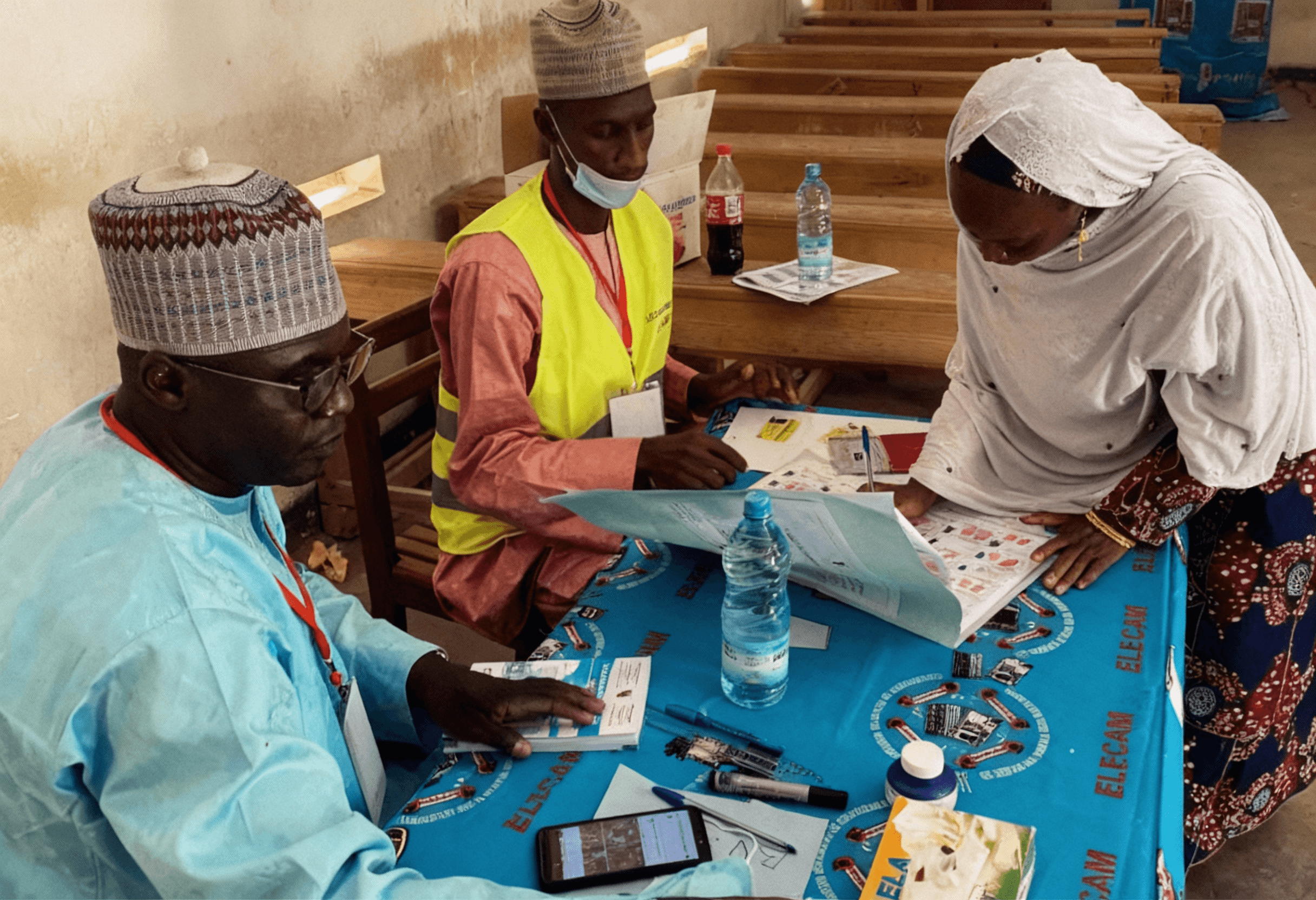 Election officials at an ELECAM office in Cameroon verify registers beside sealed transparent ballot boxes; staff work on laptops under bright fluorescent lights.
