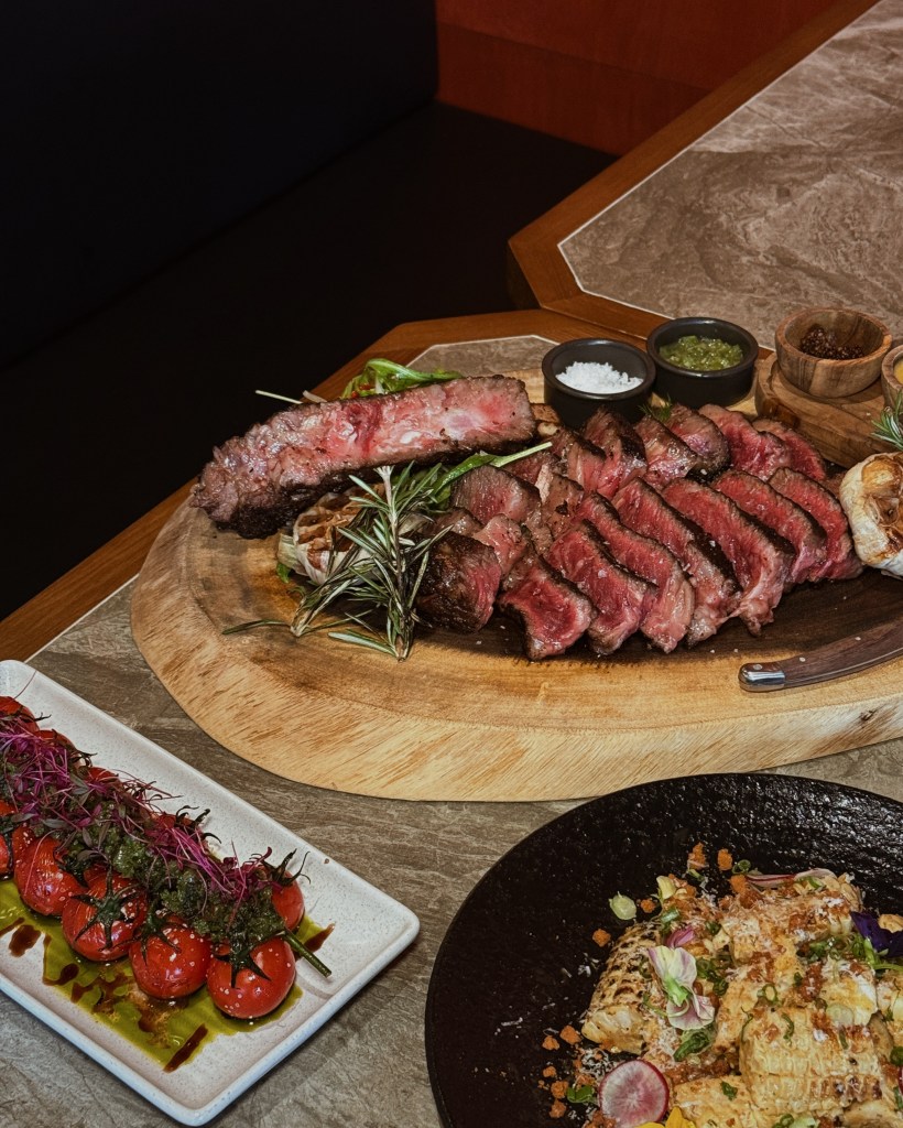 A beautifully presented platter featuring a large portion of perfectly grilled tomahawk steak, sliced and garnished with herbs. Accompanied by colorful appetizers including roasted tomatoes drizzled with olive oil and a vibrant salad on the side.