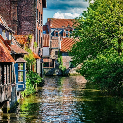 Kanal okružen zgradama od opeke s crvenim crijepom na lijevoj strani i bujnim drvećem na desnoj strani, pod djelomično oblačnim nebom.