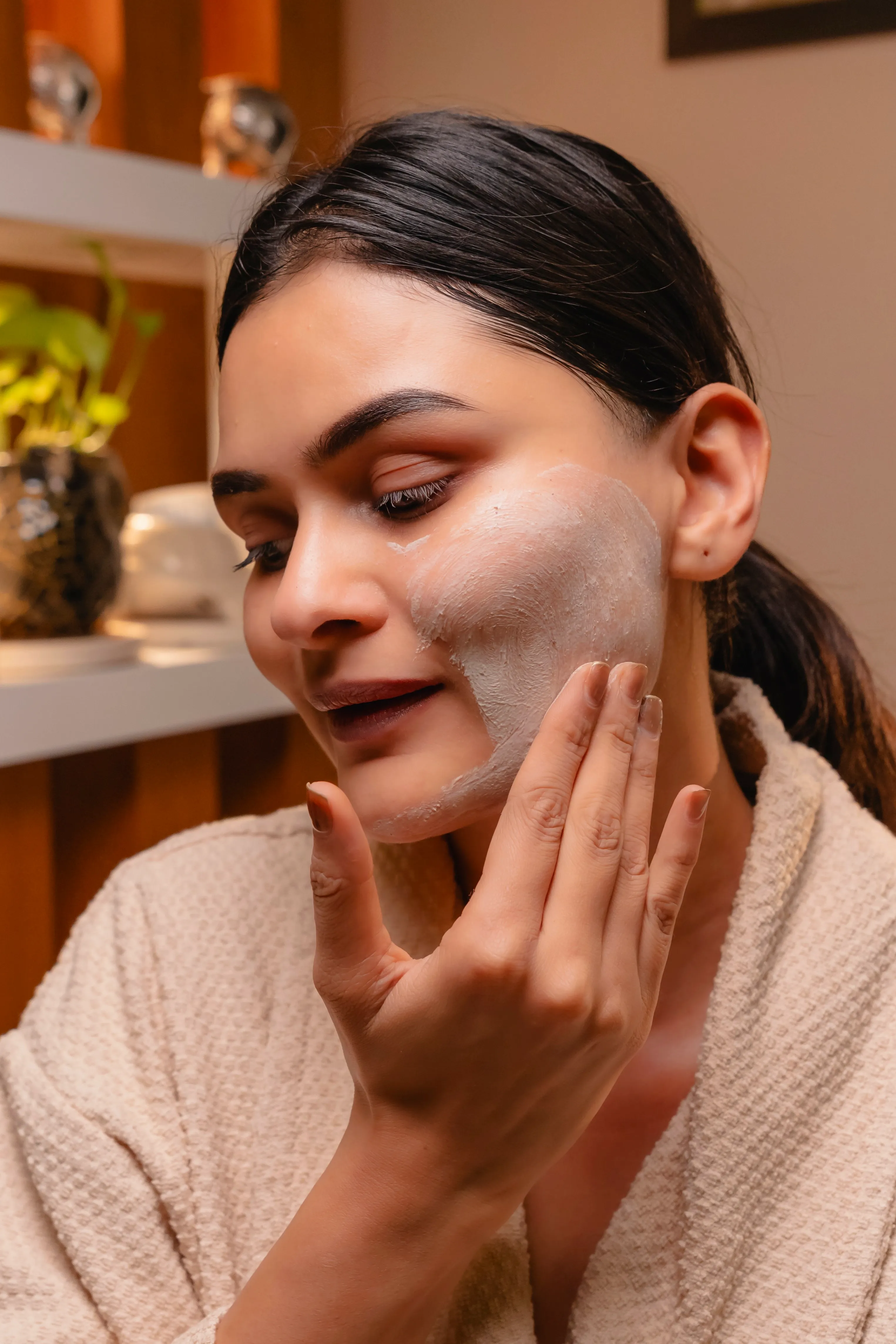 A woman applies a white face mask, smiling gently in a cozy, well-decorated environment.