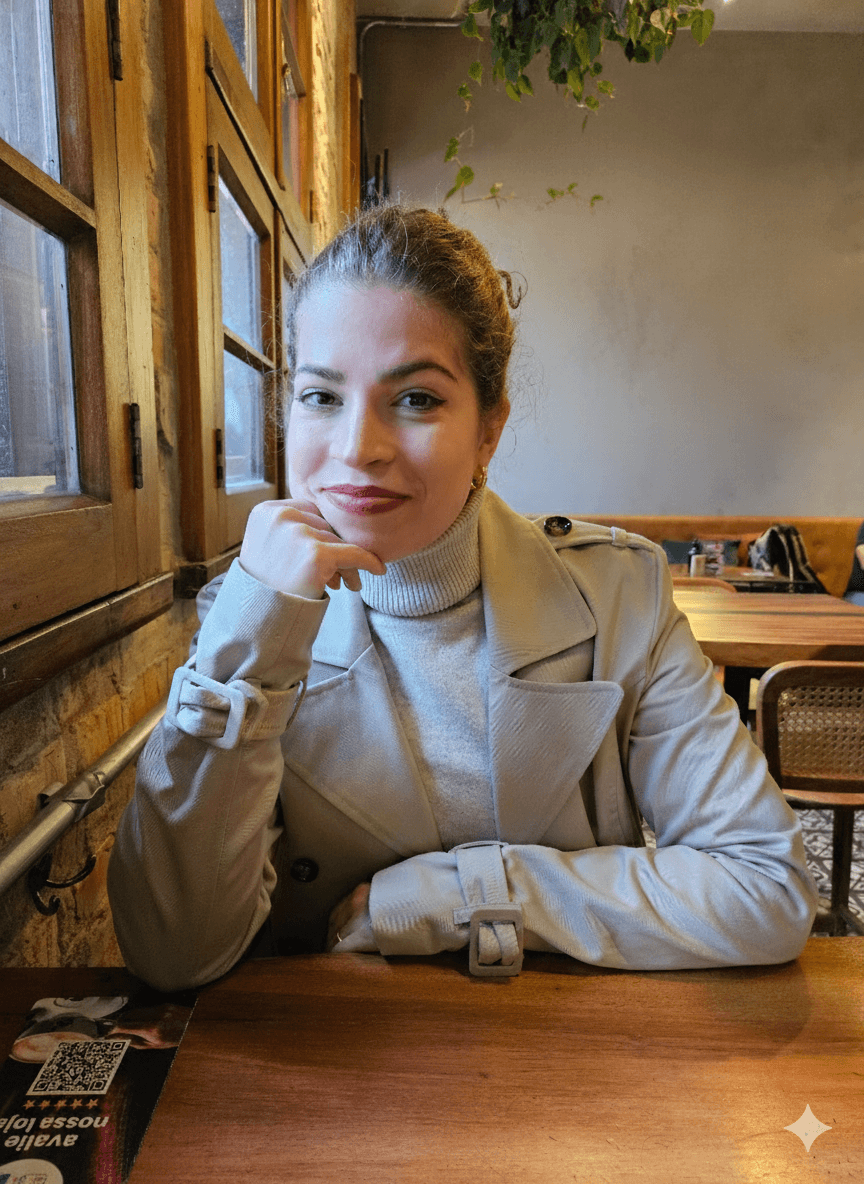 A person with curly hair and glasses smiles at the camera, seated in a cozy workspace with warm lighting.