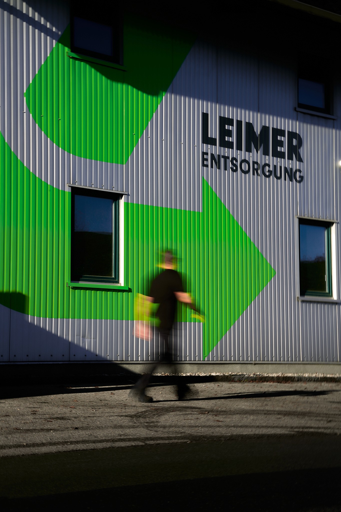 Person geht an einem Gebäude mit grünen Recyclingpfeil und der Aufschrift "Leimer Entsorgung" vorbei.