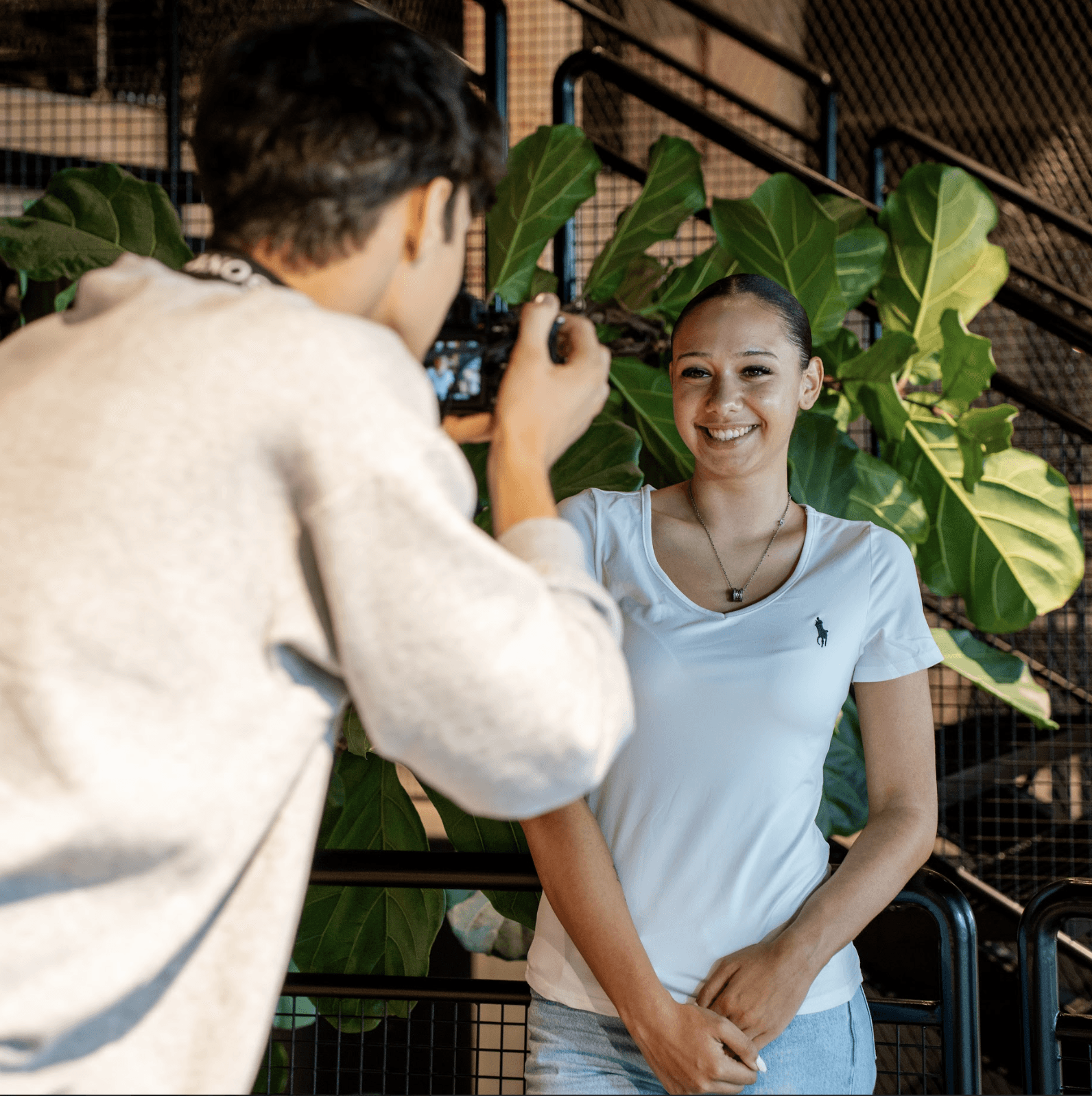 A young person with curly hair smiles at the camera while sitting in front of a laptop against a dark background.