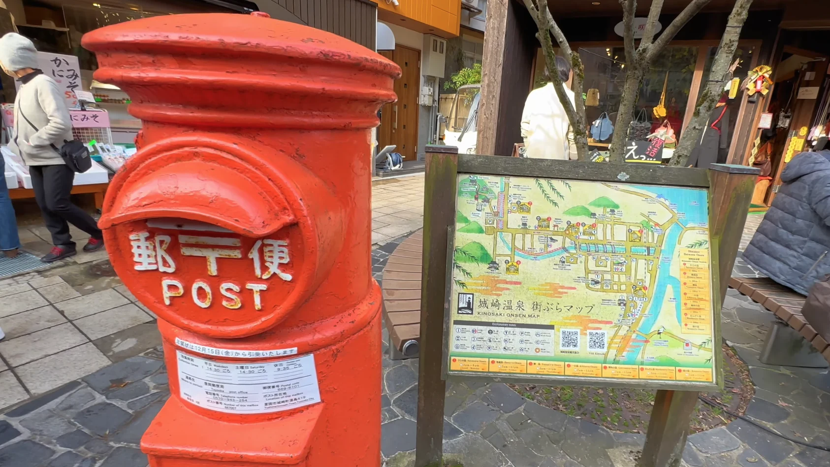 A red Japanese post box standing beside an illustrated Kinosaki town map board.
