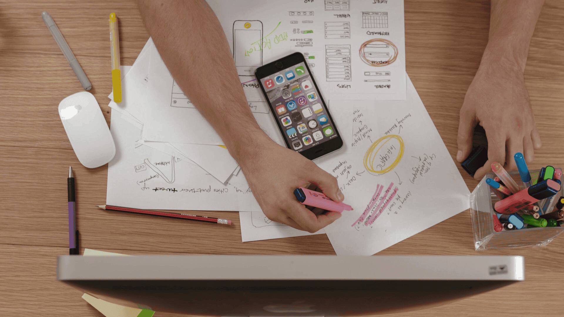 A person is working at a desk. They are using a marker to draw on a piece of paper with sketches and diagrams. There are other pieces of paper, a phone, a computer monitor, a mouse, and a variety of pens and pencils on the desk.