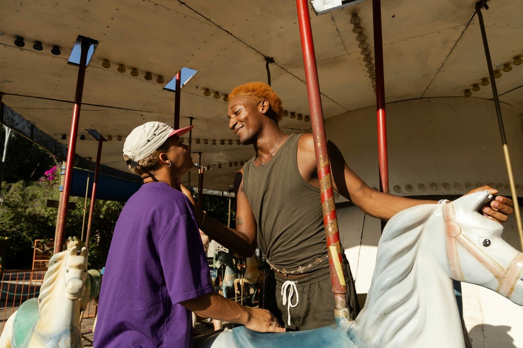 Two young men sharing a lighthearted moment on an old-fashioned carousel, one with bleached hair and a tank top, the other in a purple shirt and cap, enjoying a sunny day at an amusement park.