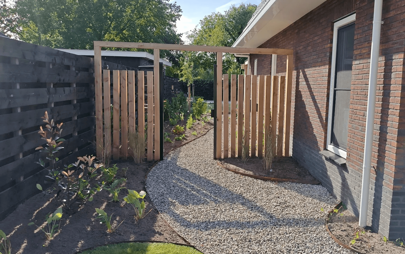 Kindvriendelijke tuin met een gebogen grindpad, houten poort, jonge beplanting en een zwarte schutting naast een nieuwbouwwoning in Friesland.