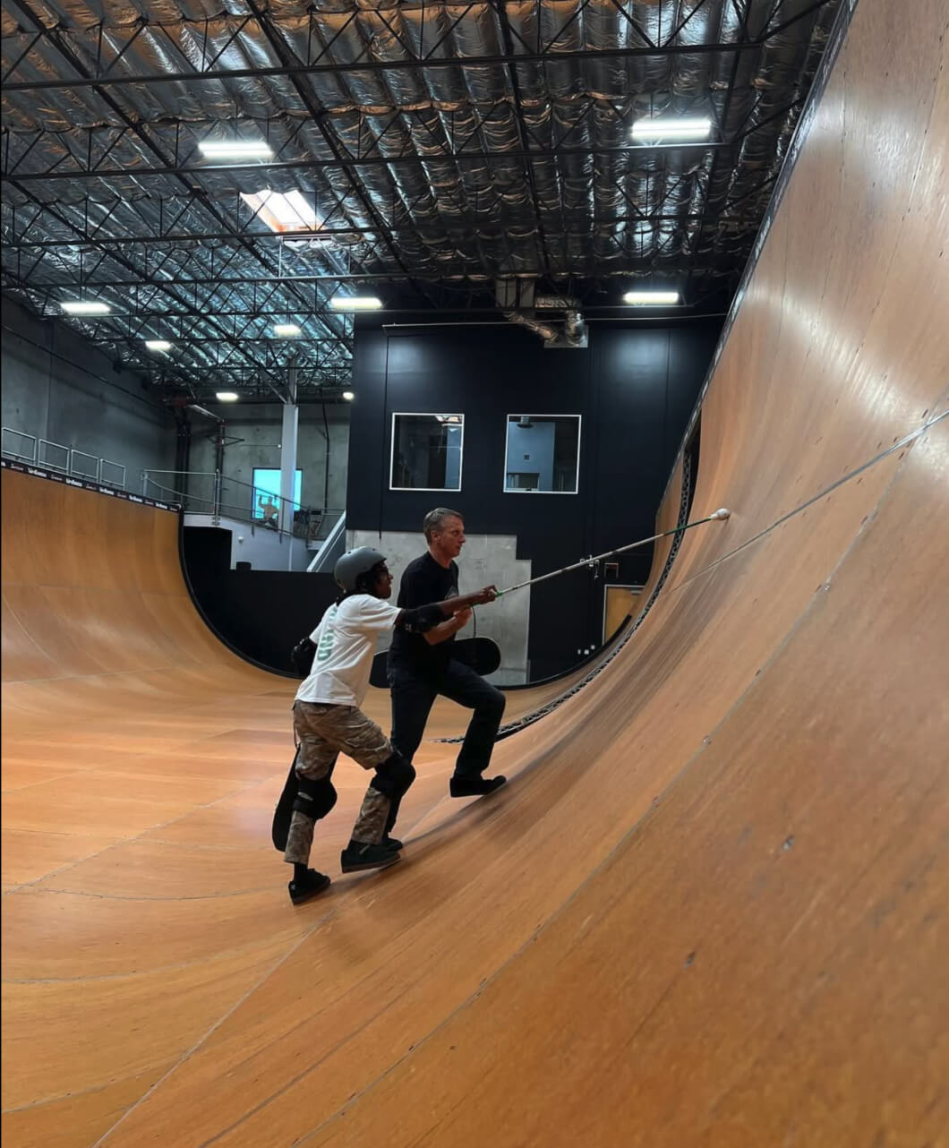 Photo of Coco with legendary pro skateboarder Tony hawk in the half pipe at tony’s house. Tony is guiding coco, showing him where the seams are in the ramp. both Coco and Tony each have a hand on Coco’s cane, feeling around the upper section of the halfpipe