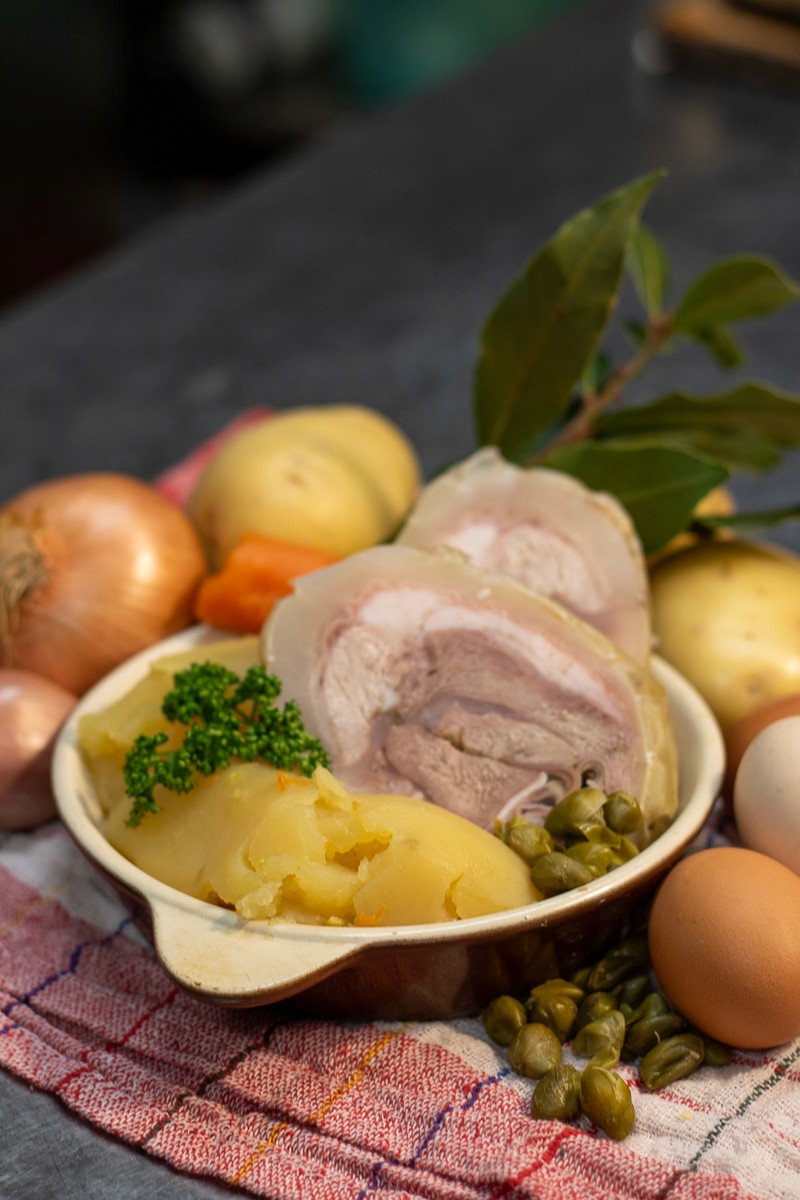 Person with a mustache wearing a black t-shirt carrying a tall stack of pizza boxes inside a dimly lit bar or restaurant.