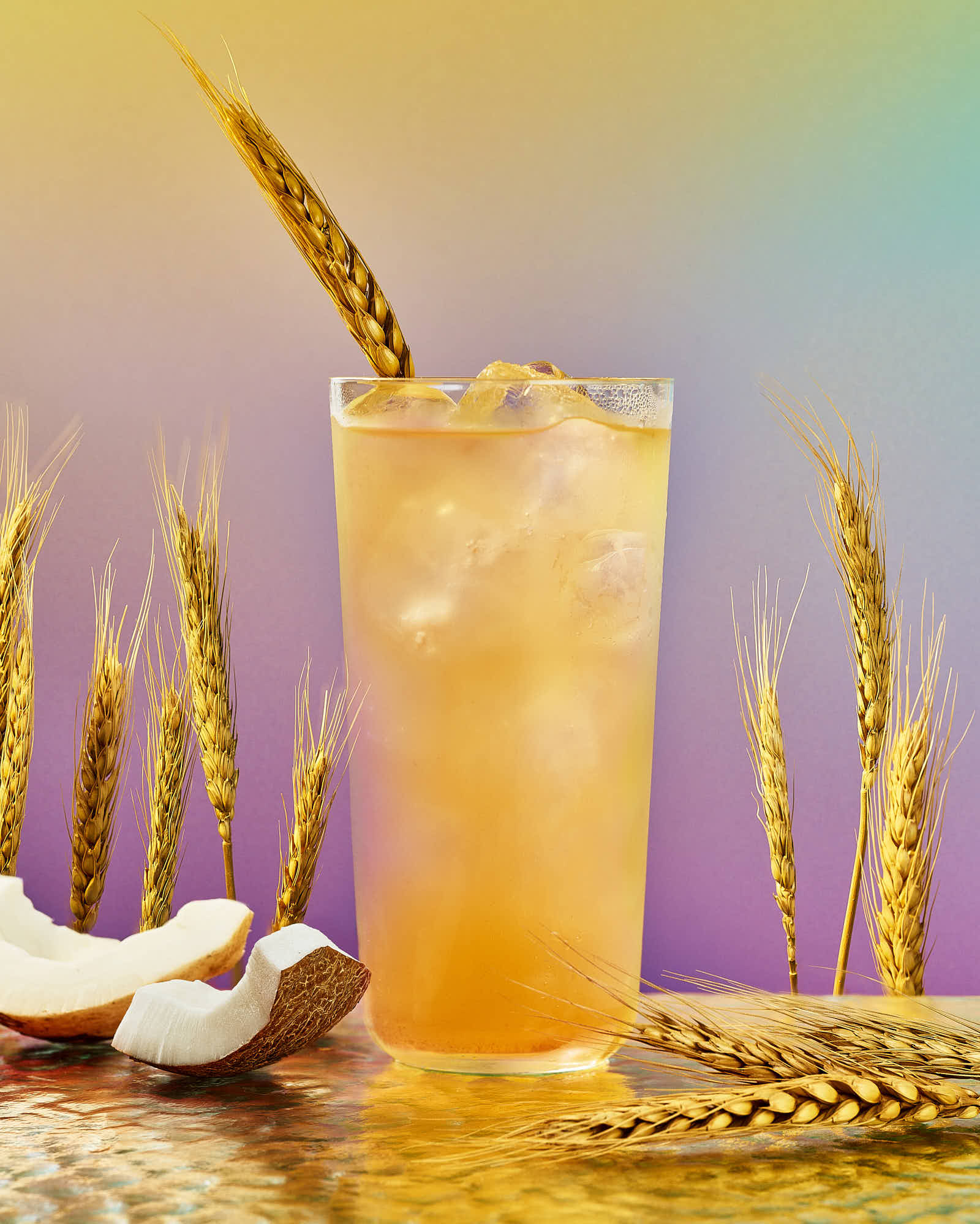 A tall glass filled with a vibrant orange drink, garnished with a straw and a piece of wheat, against a colorful background.