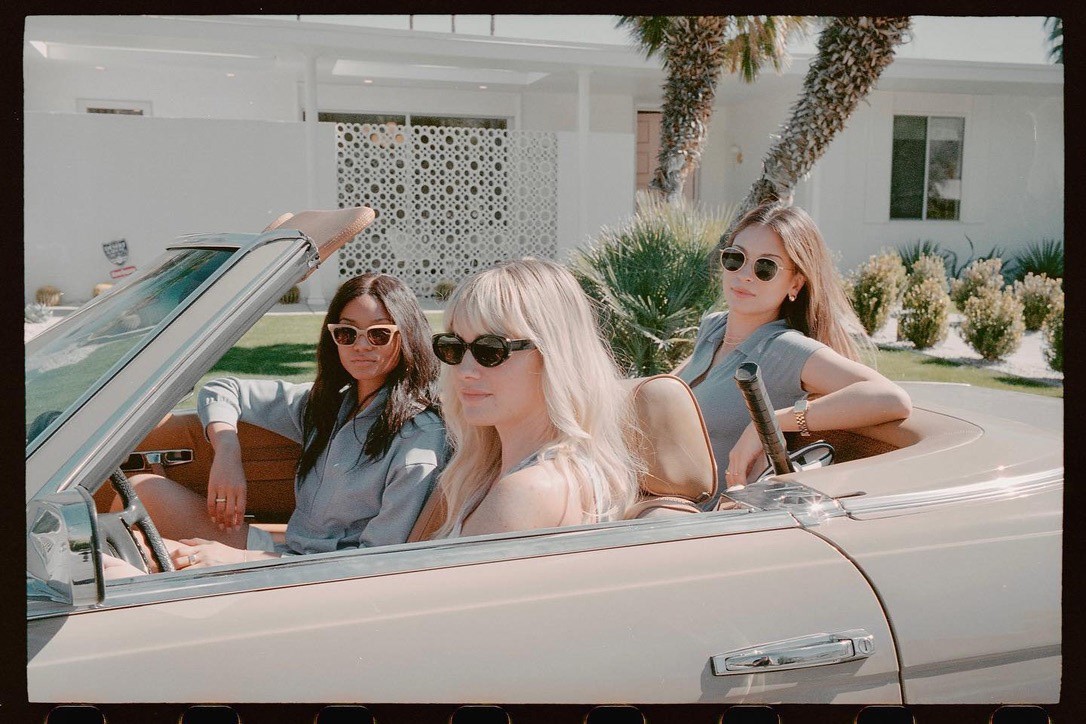 Three women in sunglasses riding a vintage convertible, sunlit