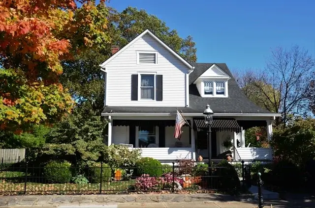 Traditional white house with a front porch, autumn trees, and a fenced garden.