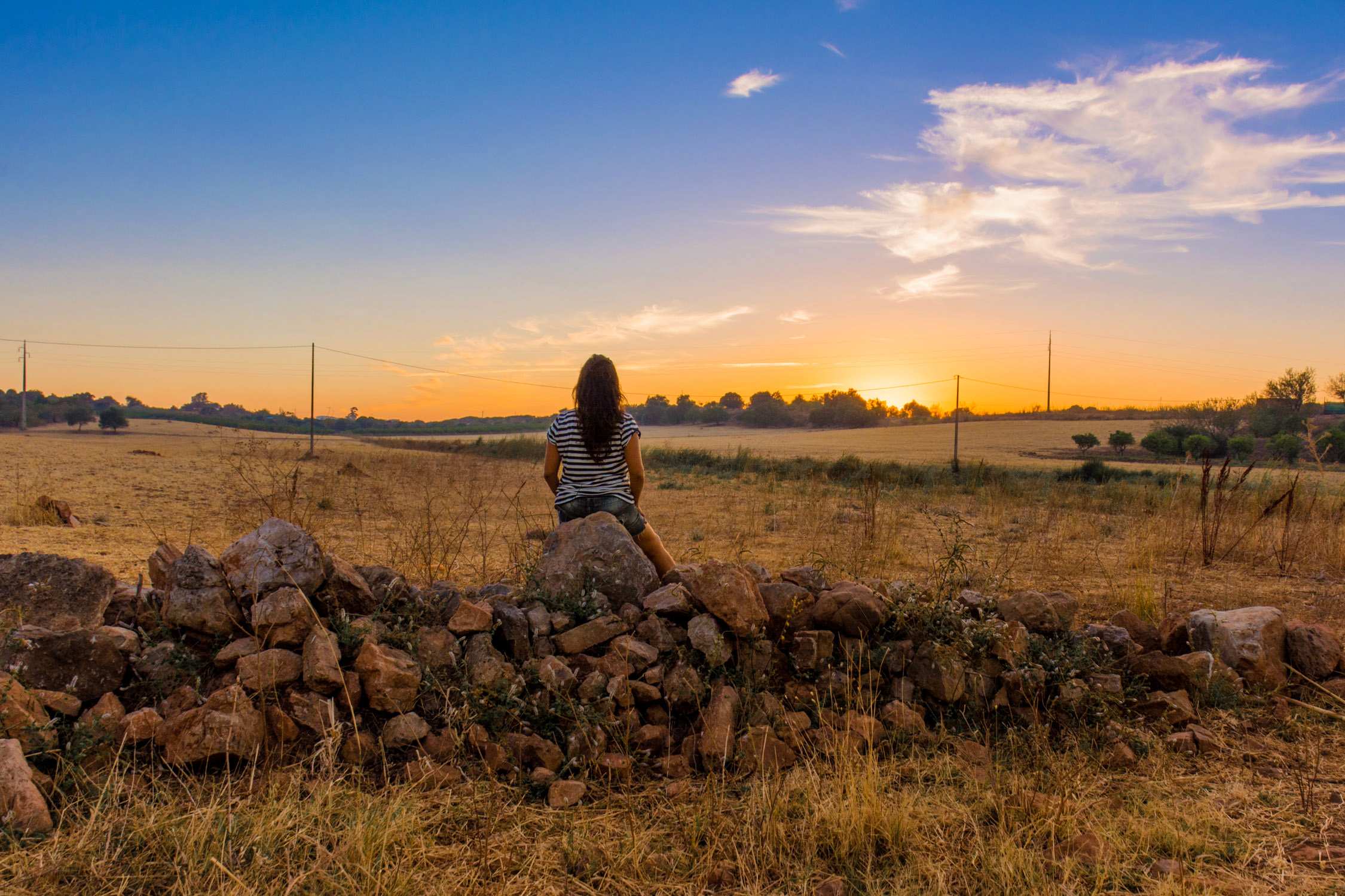 A person sits on a low stone wall in a rural field at sunset, looking toward a golden horizon under a blue sky.