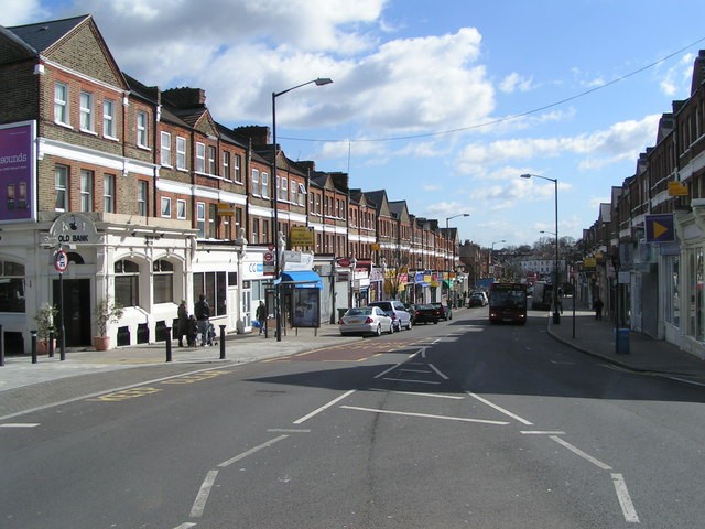 A smiling man with curly hair is sitting at a desk, looking at the camera, with a laptop and a lamp in the background.