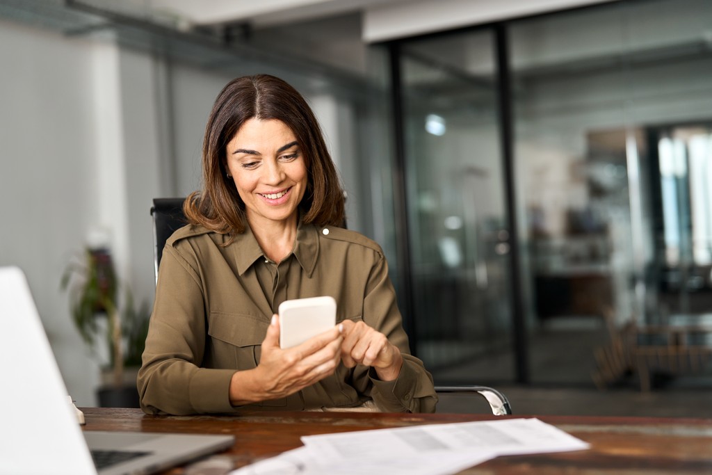 woman in a green button-up shirt smiles while looking at her phone at a desk in a modern office, with a laptop open beside her.