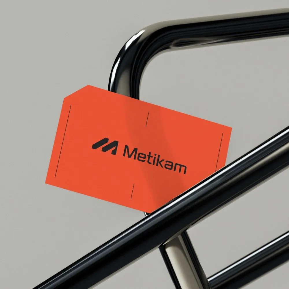 Orange Metikam business card with clipped corner resting on chrome steel tubes, stationery design