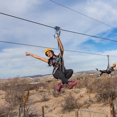 Двама души се спускат по въже над суров, пустинен пейзаж, носейки каски и предпазни колани под частично облачно небе.