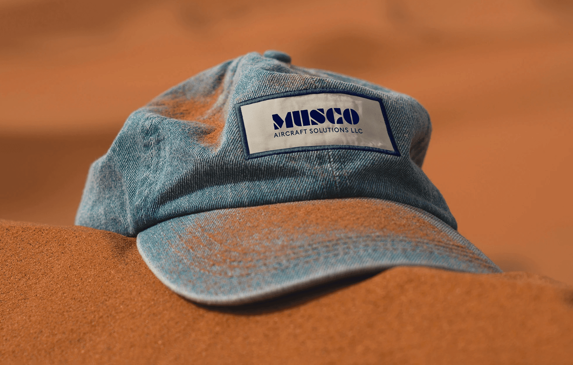 Graphic design mockup of a hat in the sand for Musco Aircraft Solutions — modern, geometric wordmark logo with a clean brandmark with a clean sans serif typography, designed by Will Lefkovich.