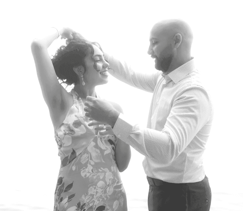 Black and white photo of a man in a button-up shirt holding hands with a woman in a dress, smiling at each other in a joyful and elegant moment.