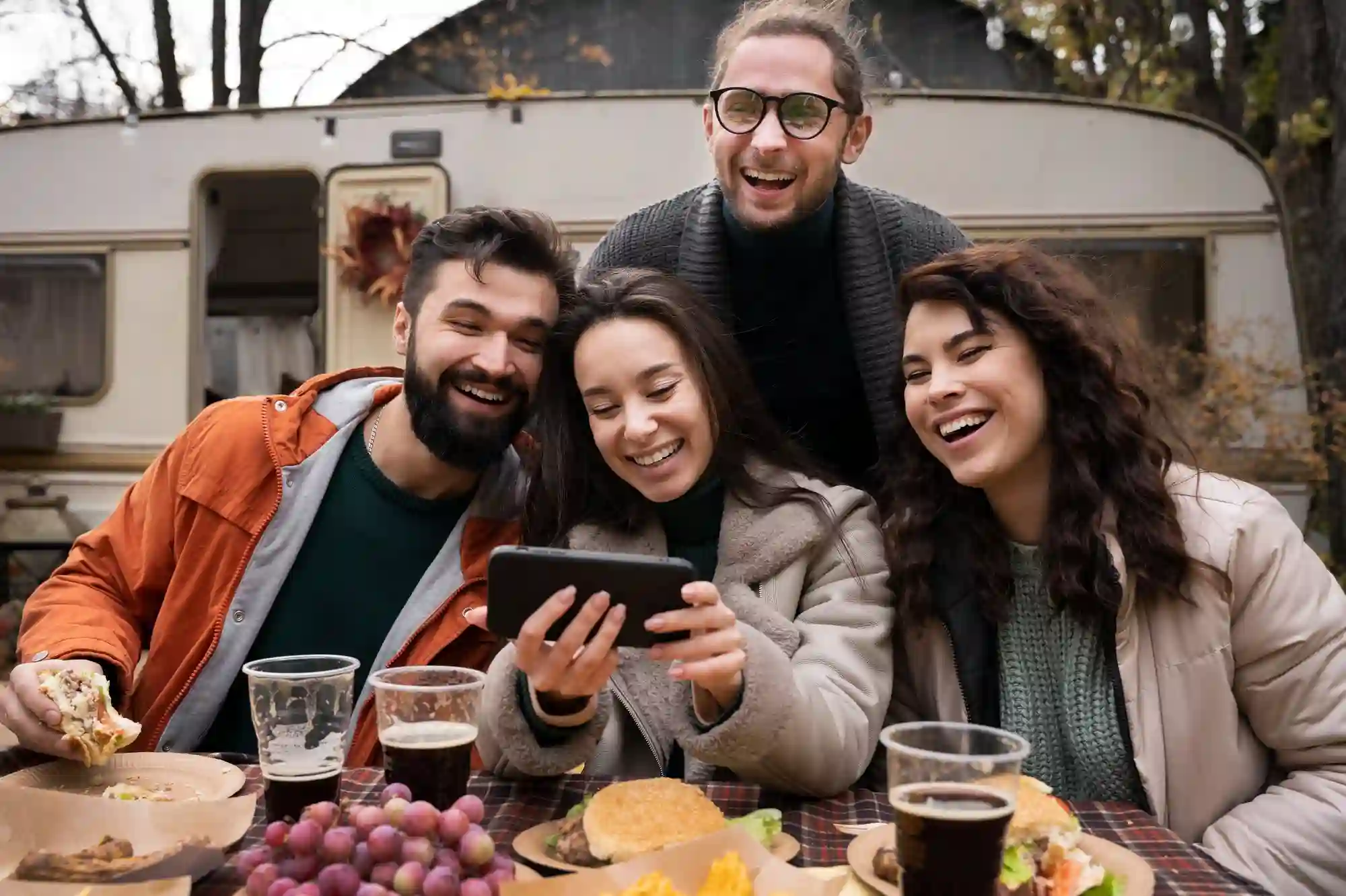 Four friends laugh and share a meal outdoors while looking at a smartphone during a cozy autumn camping trip.