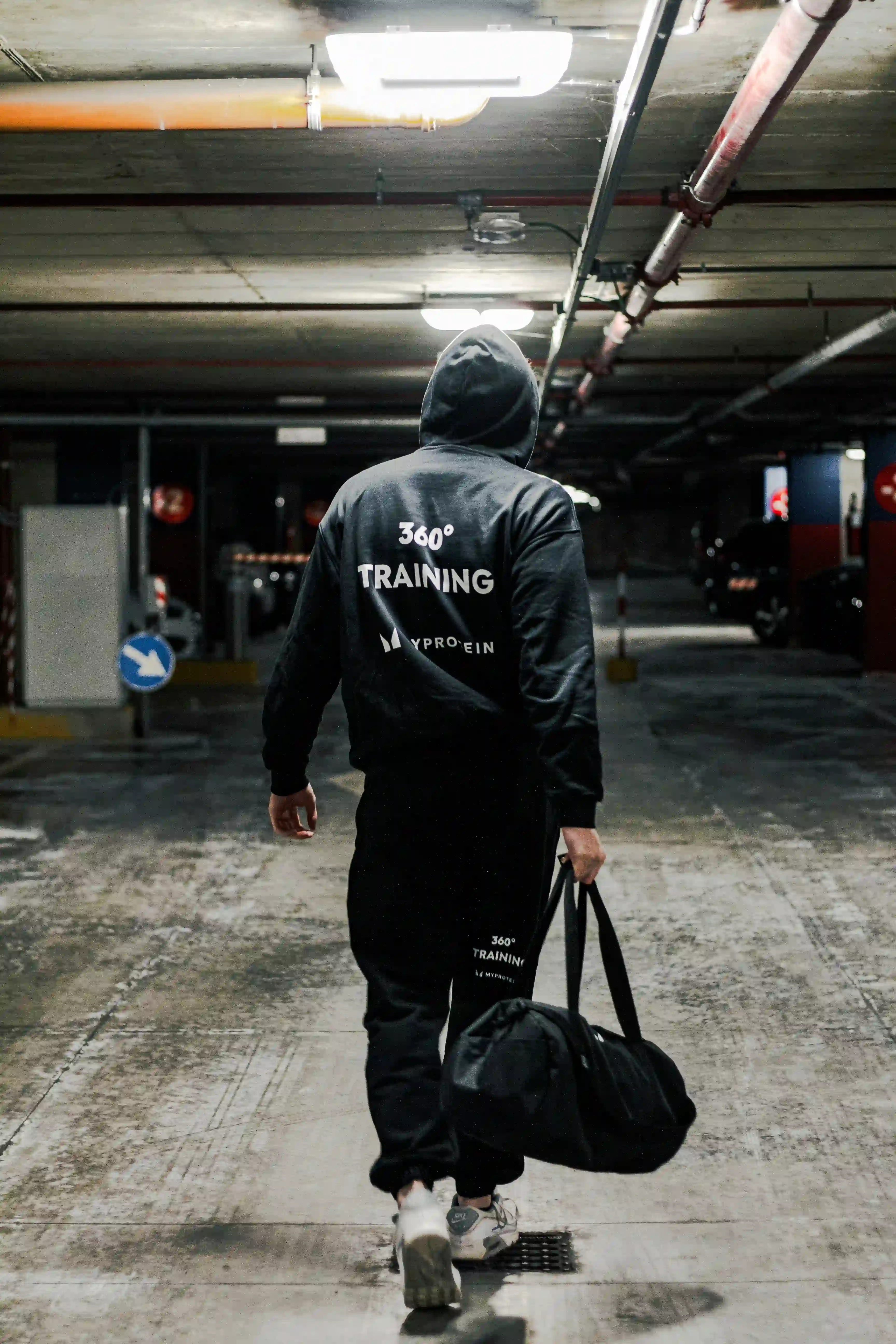 Person wearing a black hoodie and carrying a gym bag while walking through an underground parking garage.