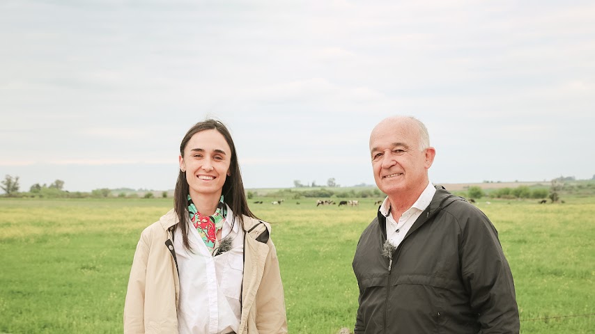 A woman and a man stand smiling in a lush green field with a cloudy sky. 