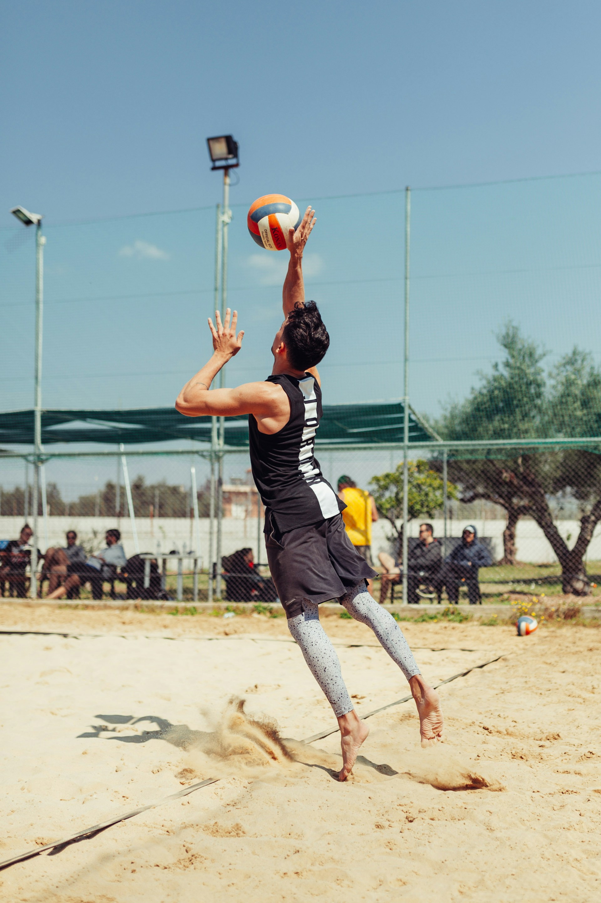 A beach volleyball player in a black tank top jumping to set the ball overhead on a sand court.