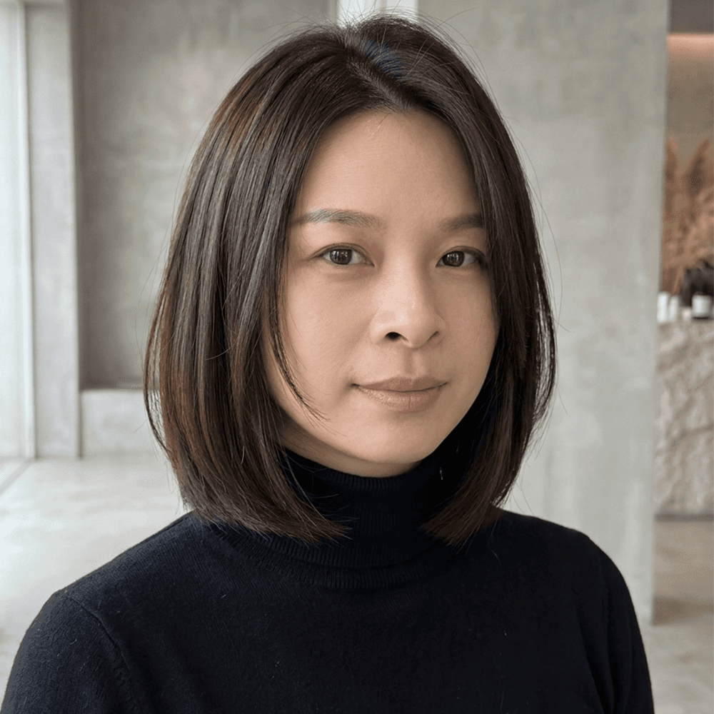 A medium close-up, professionally styled portrait of a woman with dark brown, sleek, chin-length bob-cut hair with a center part and face-framing layers. She wears a simple black knit turtleneck and looks calmly at the camera with a subtle smile. She is in a minimal, contemporary interior setting with a blurred background featuring natural light, textured grey concrete walls, large windows, and dried pampas grass.