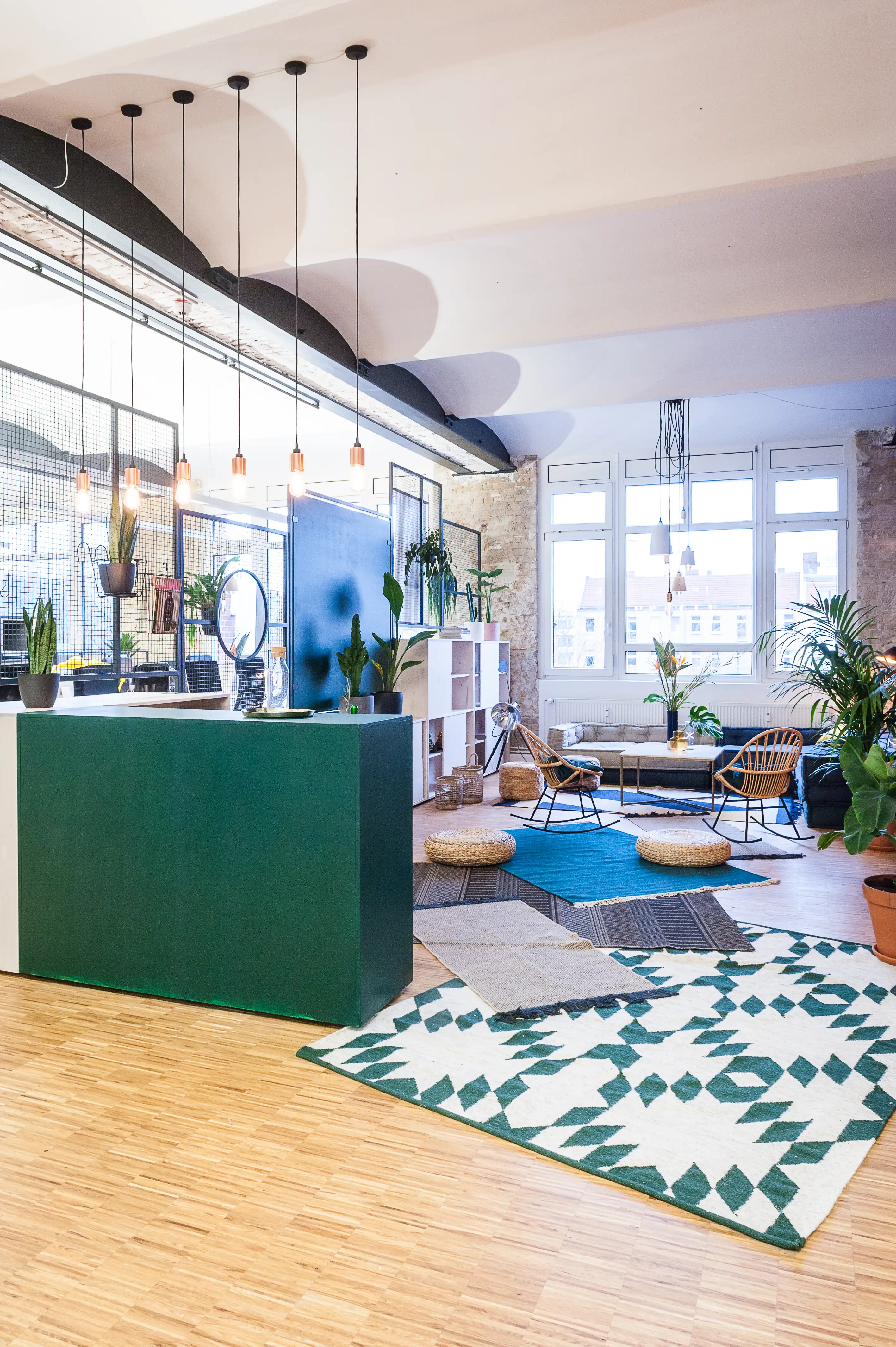Reception of INTERMATE x TRUEMATES with a dark green counter, copper pendant lights, scalloped black ceiling frieze and a lounge area with patterned green-and-white rugs and rattan chairs.