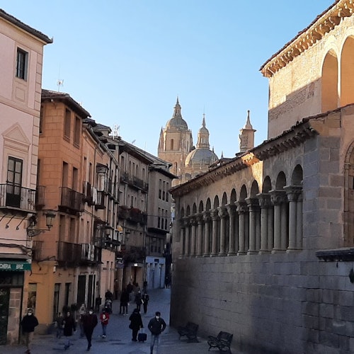 A narrow street lined with historic buildings leads to a cathedral with multiple domes. People walk down the street.
