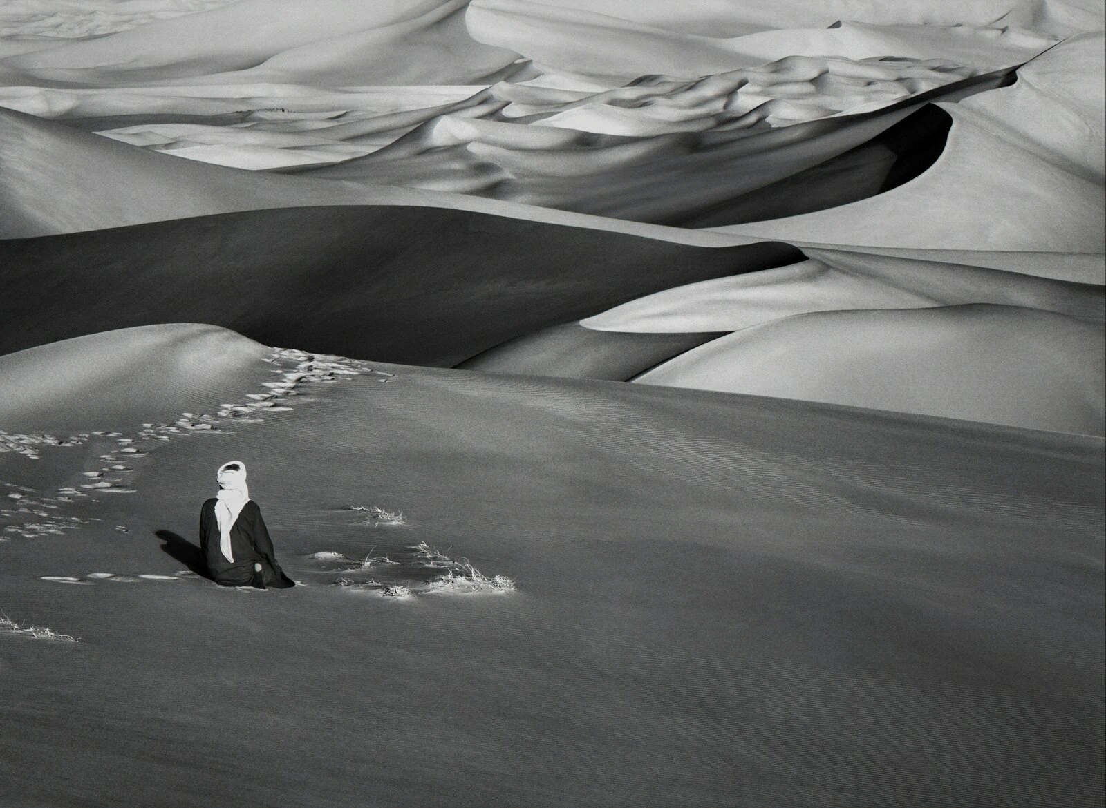 A solitary figure in dark robes and white headscarf seated on a smooth dune slope, a trail of footprints leading behind them, towering dunes receding into the distance, black and white