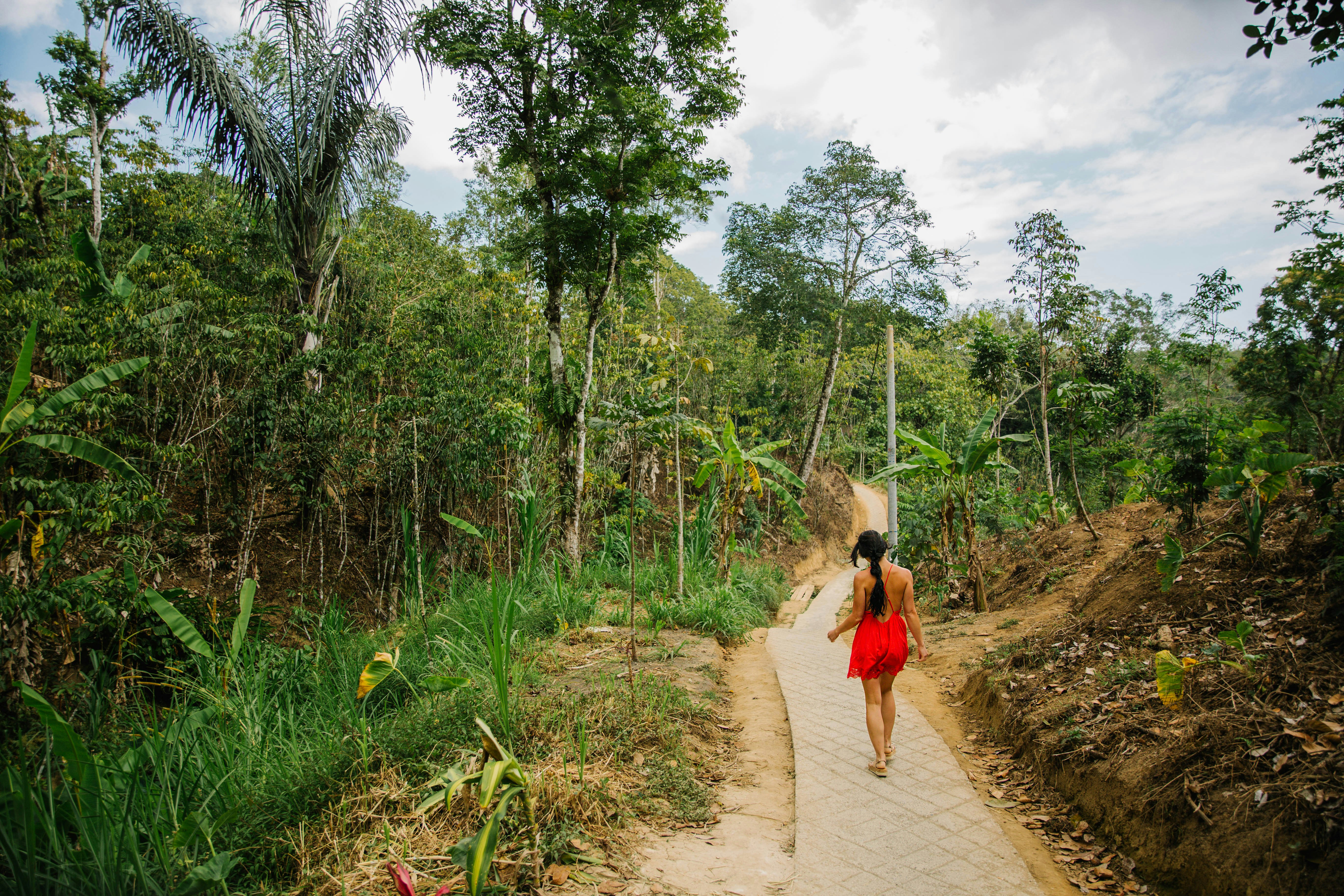 woman in red dress walking on pathway between green grass and trees during daytime