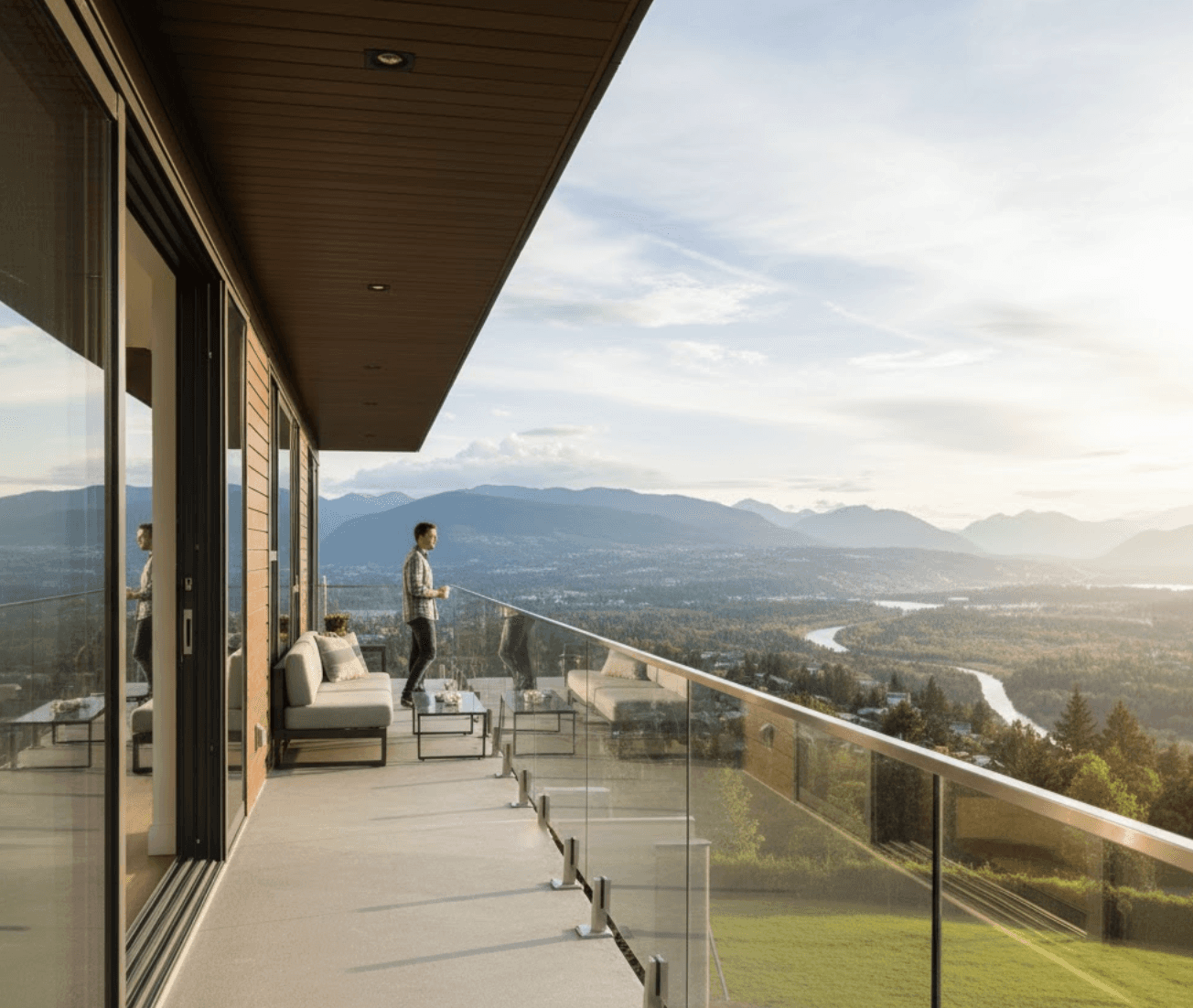 A modern home balcony with glass railings, offering an unobstructed panoramic view of a scenic valley with mountains and a river, typical of the Vancouver area. A person is standing on the balcony, enjoying the view, emphasizing the clear sightlines provided by the glass. The image should highlight the aesthetic appeal and functionality of glass railings in preserving valuable landscapes.