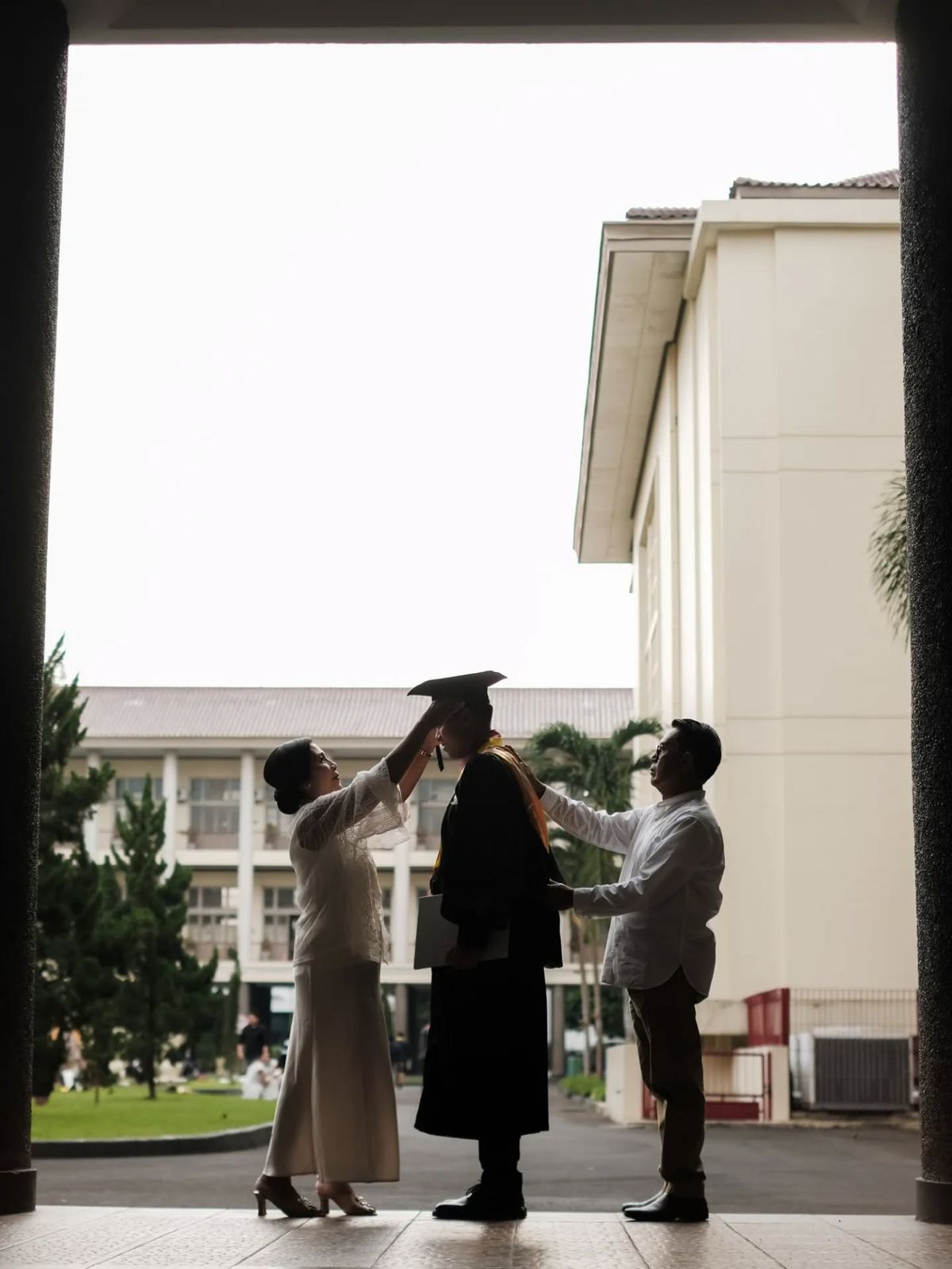 Graduate standing outdoors while two family members adjust their cap and sash