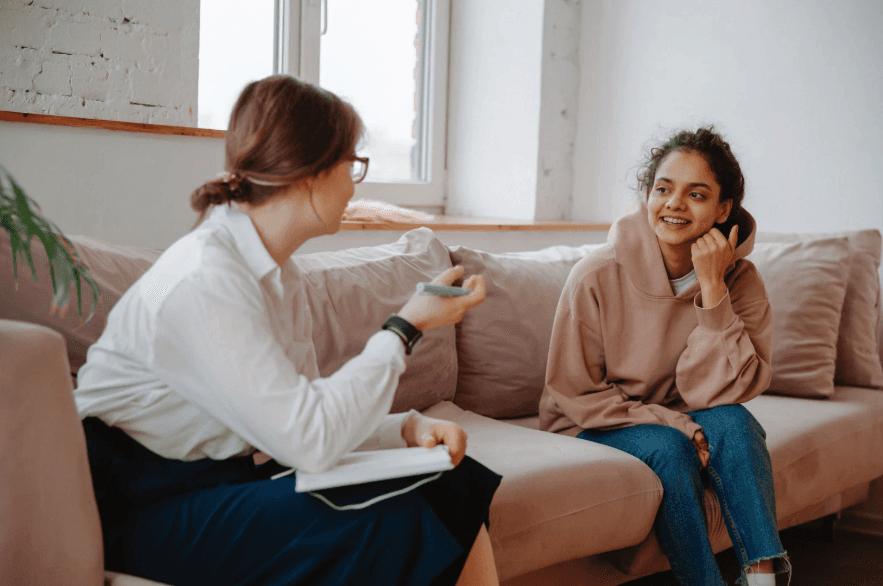 A cozy and inviting spa room with a neatly made bed, soft lighting, and warm wooden tones. The scene is partially framed by green plants, adding a natural and soothing touch.