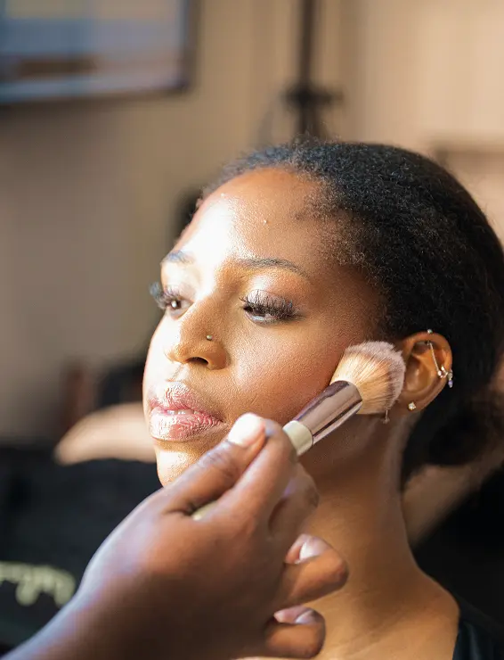 A person receives makeup applied with a brush, highlighting their cheek. The lighting is warm, creating a focused and calm atmosphere.