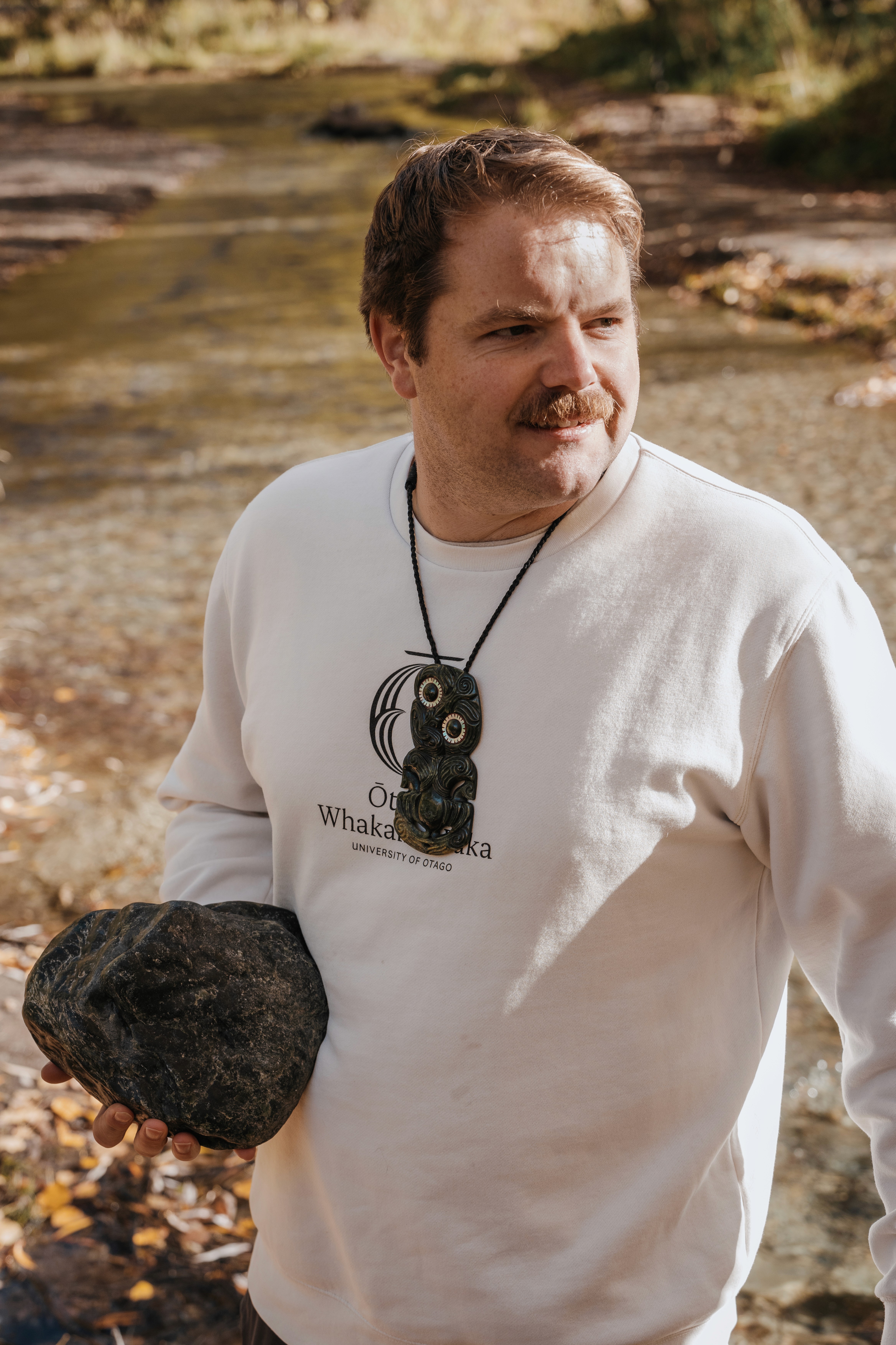Brendon beside an awa, holding a stone and wearing a pounamu pendant.