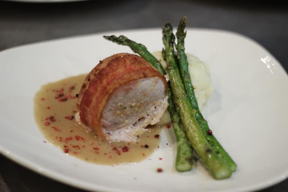 Bacon wrapped pork tenderloin plated with creamy sauce, asparagus, and mashed potatoes on a white plate.