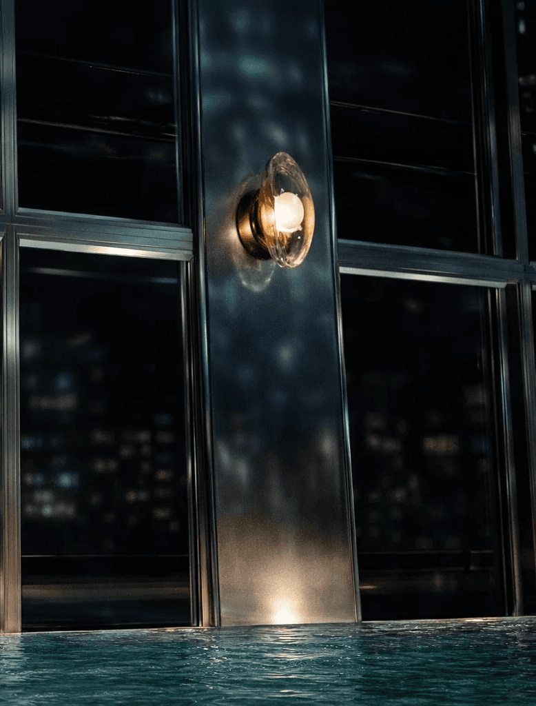 Luxury bronze and glass wall sconces illuminating a modern indoor pool area with floor-to-ceiling windows and night city view reflections on the water.