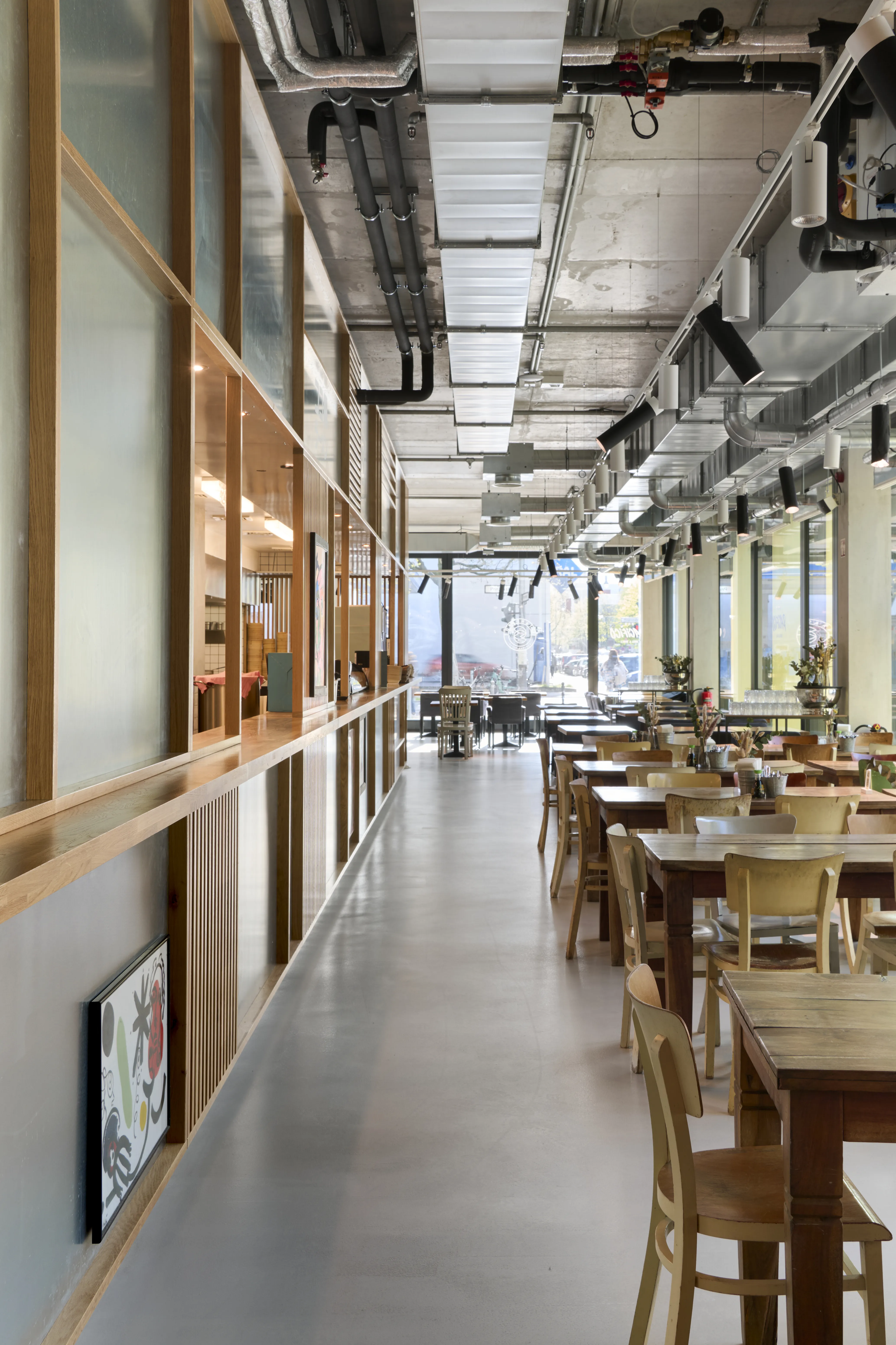 Row of wooden dining tables along a wood-framed frosted glass wall at PARKER BOWLES & FRIENDS, with exposed ceiling installations and daylight from large windows.