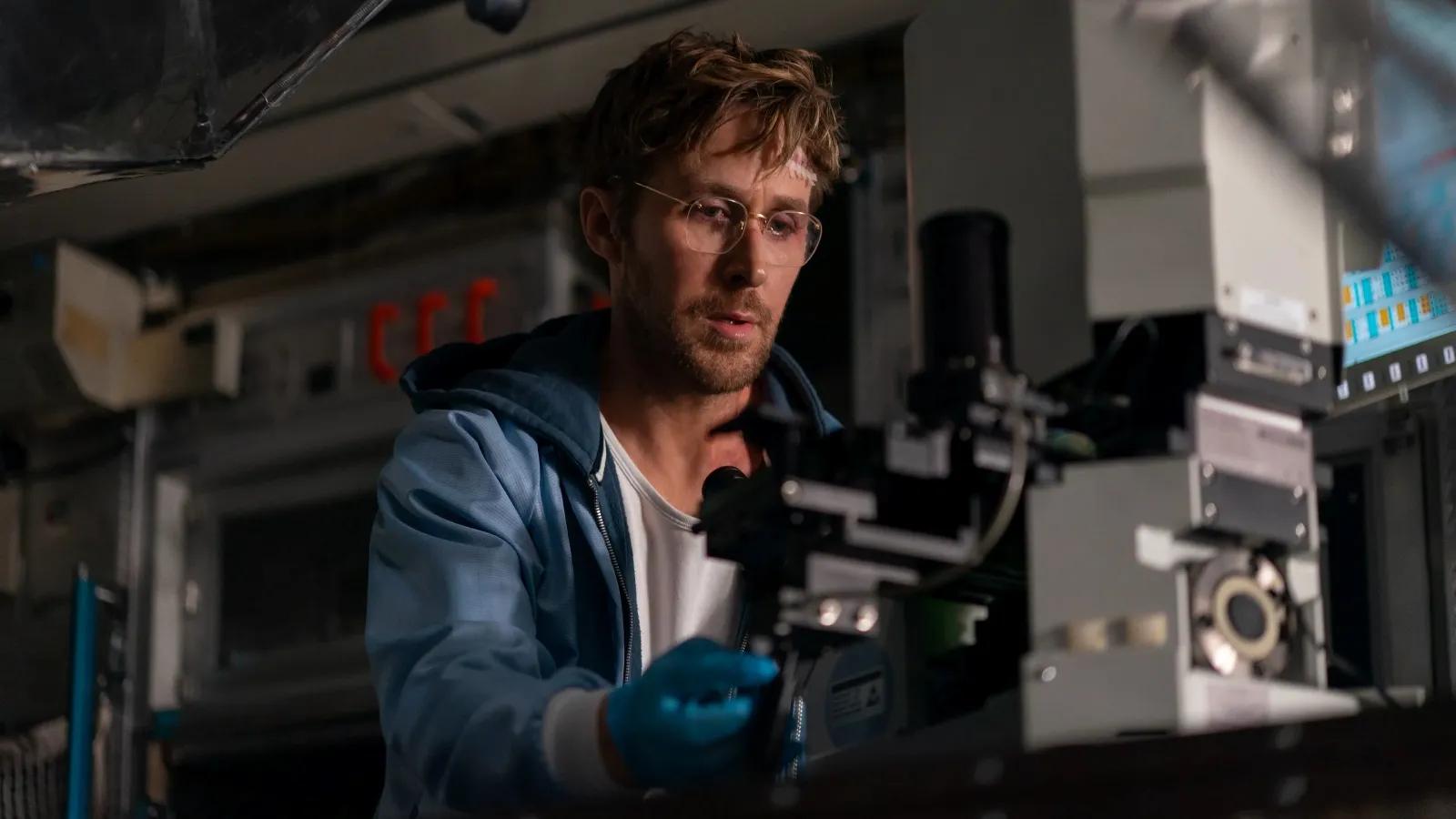 A man wearing glasses and a blue jacket meticulously examines a piece of scientific equipment in a dimly lit laboratory, focusing intently on the research task.