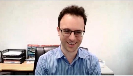 A man with glasses smiles while sitting at a desk with files and paperwork in the background.