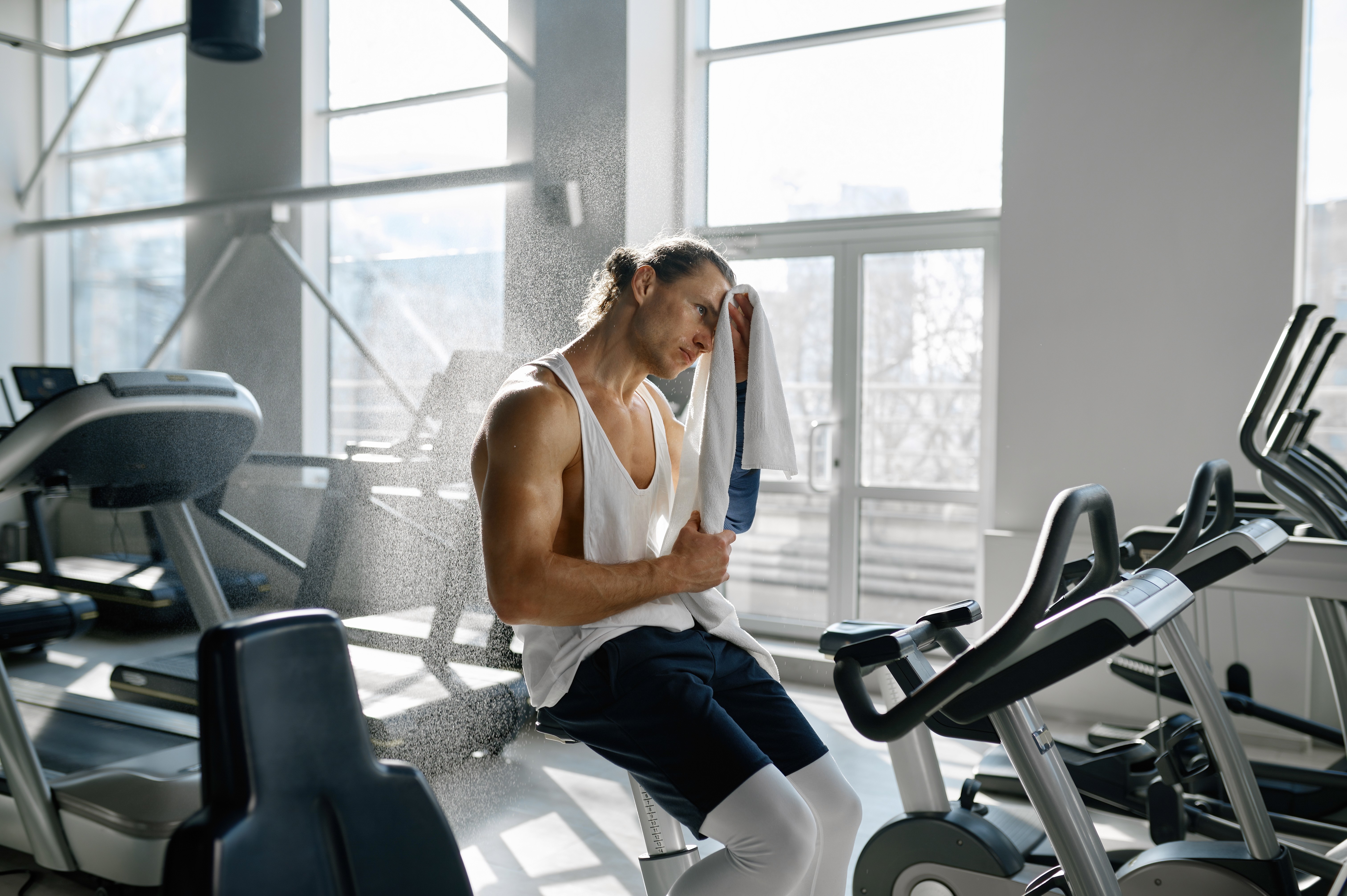 Man sitting on an exercise bike in a gym, wiping sweat from his face with a towel, taking a break after an intense workout