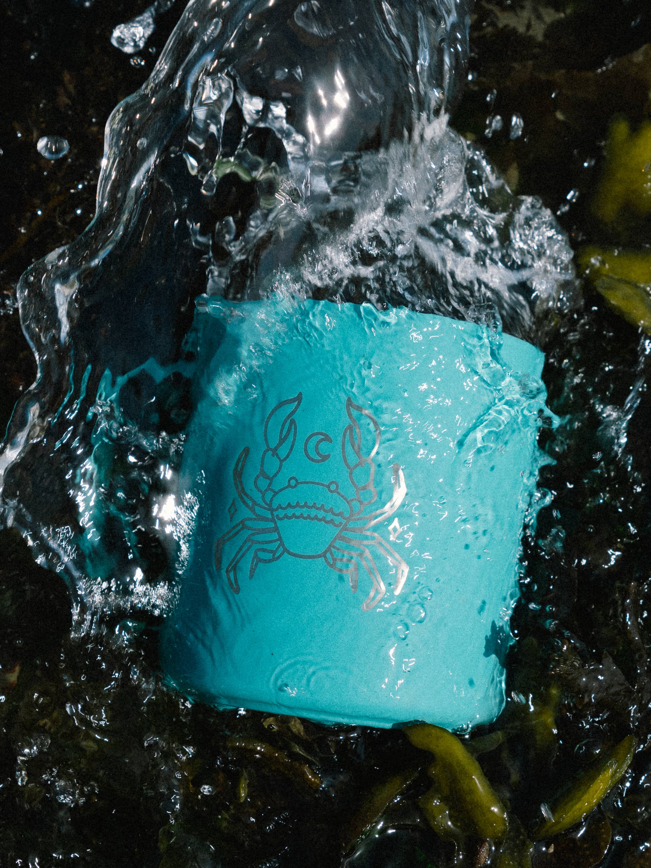 Turquoise metal mug with crab design splashed by water. Dark seaweed and foamy waves surround it, creating a fresh and dynamic oceanic vibe.