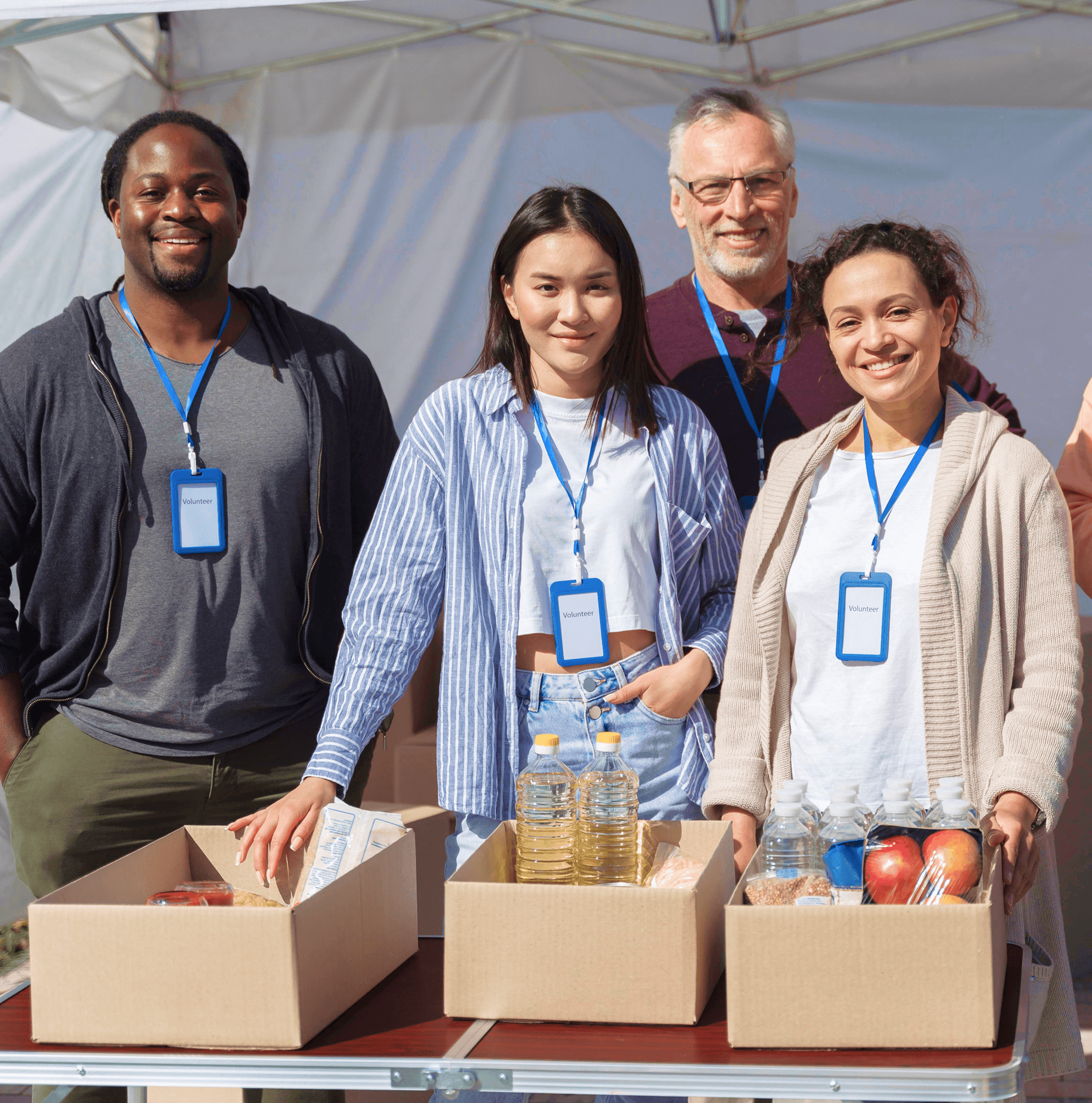 Four smiling volunteers pose together behind boxes filled with supplies at an outdoor event.